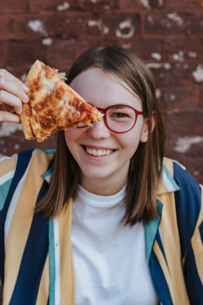 A happy teenager holding a pizza slice next to her eye, wearing red glasses, smiling outdoors.